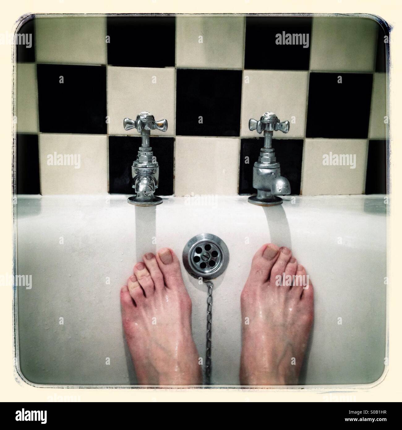 Une bonne tremper. Paire de pieds de l'homme sous une paire de robinets d'eau chaude et froide, carreaux noir et blanc arrière-plan. Banque D'Images