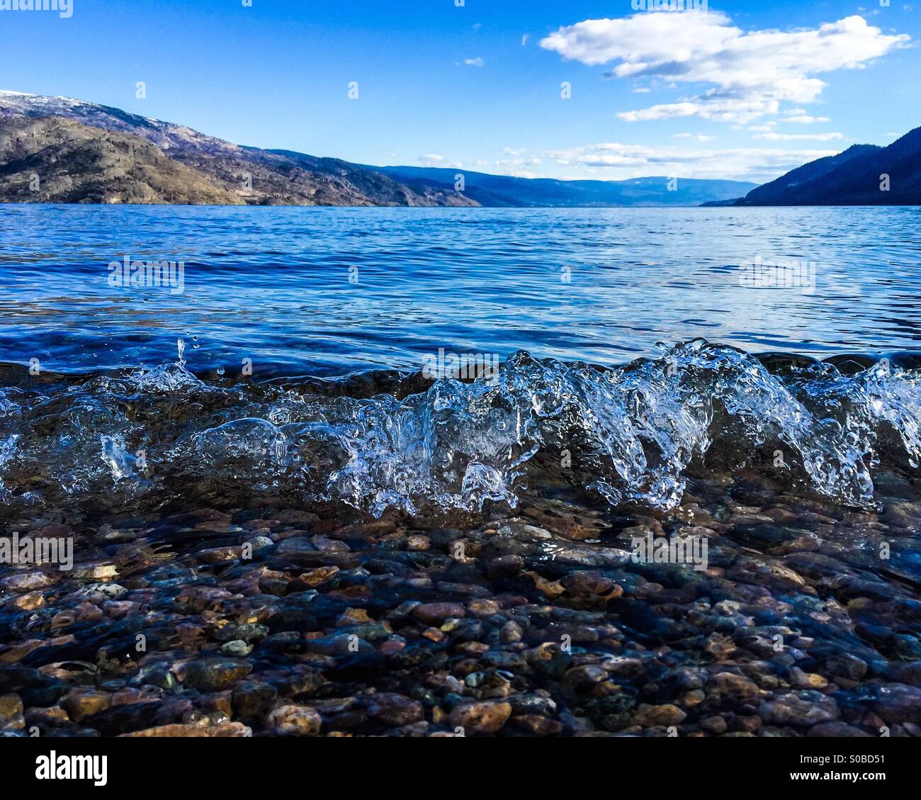 Vagues se brisant sur les rives du lac. L'eau cristalline expose le fond de galets, tandis que les montagnes en arrière-plan offrent un paysage à couper le souffle. Banque D'Images