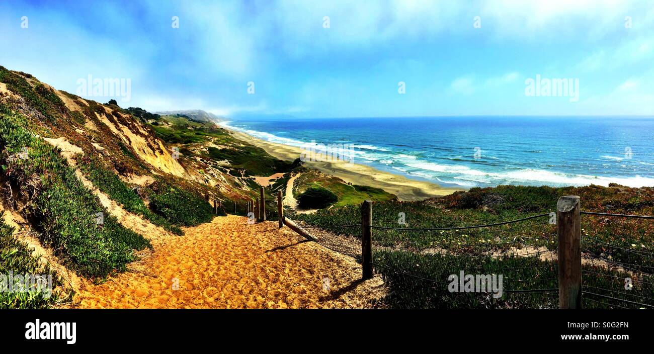 Long chemin de sable sinueux menant vers une étendue de plage apparemment infinis ci-dessous. Matin brumeux couche marine commence à peine à brûler. Fort Funston, Californie, USA. - Image de stock capturée avec un smartphone