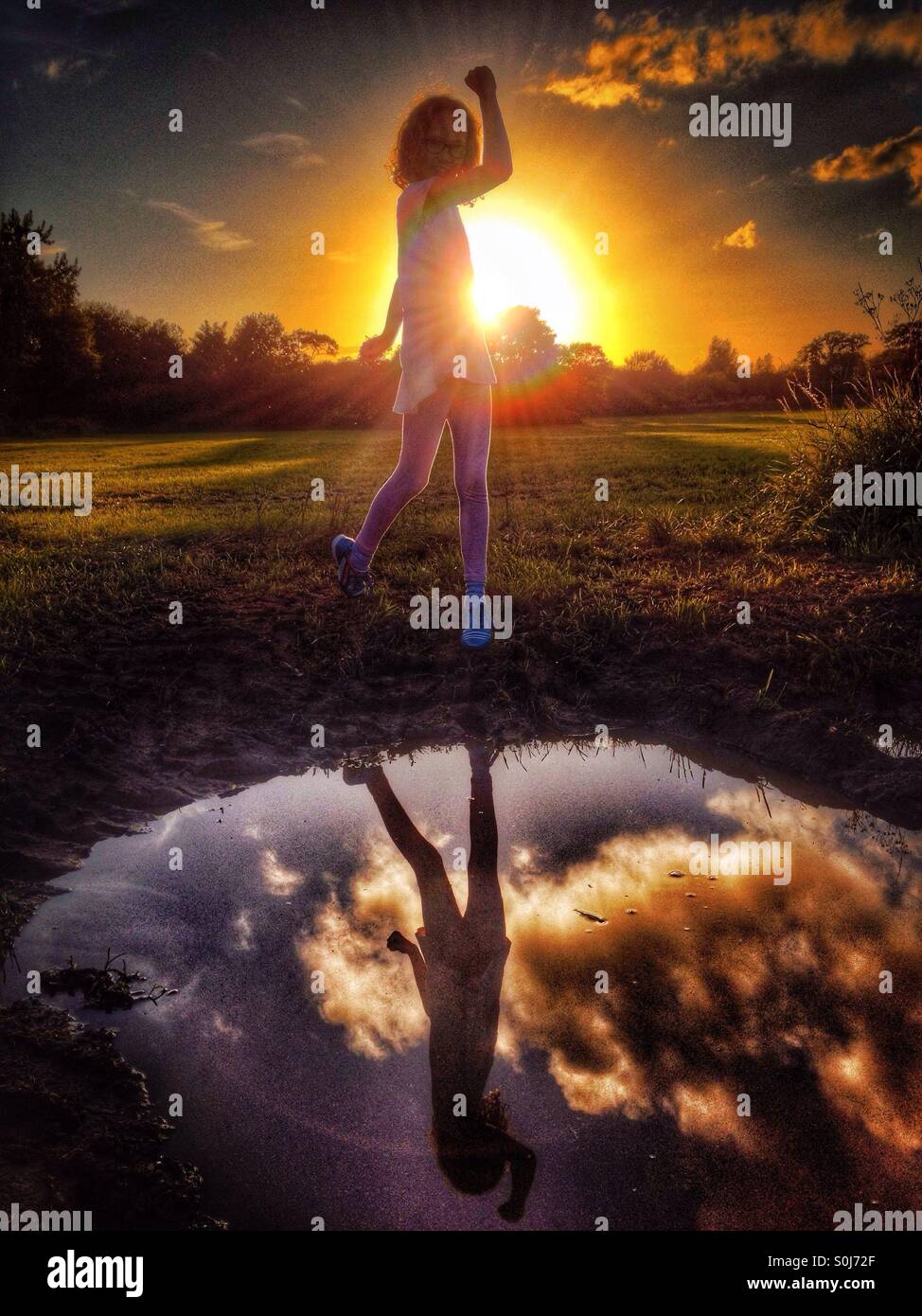 Jeune fille posant avec bras soulevé face au soleil couchant reflété in puddle Banque D'Images