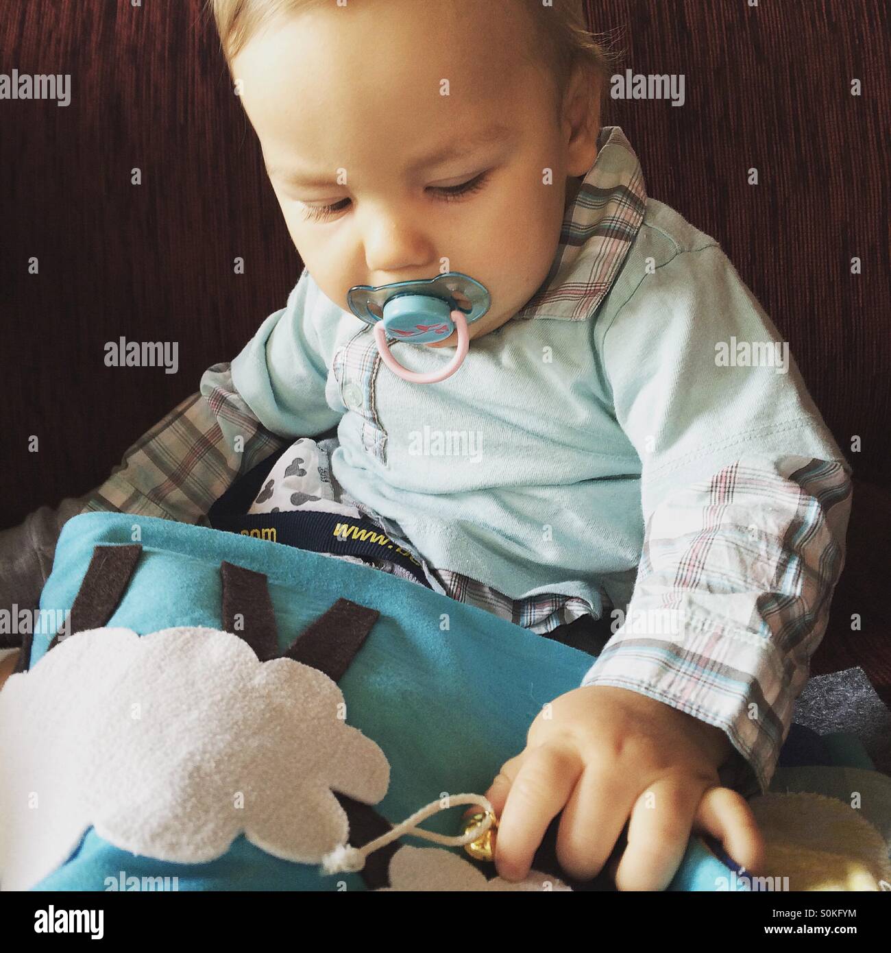 9 mois bébé garçon avec une sucette dans sa bouche, assise sur un canapé rouge est de toucher et de jouer avec un livre sensoriel Banque D'Images