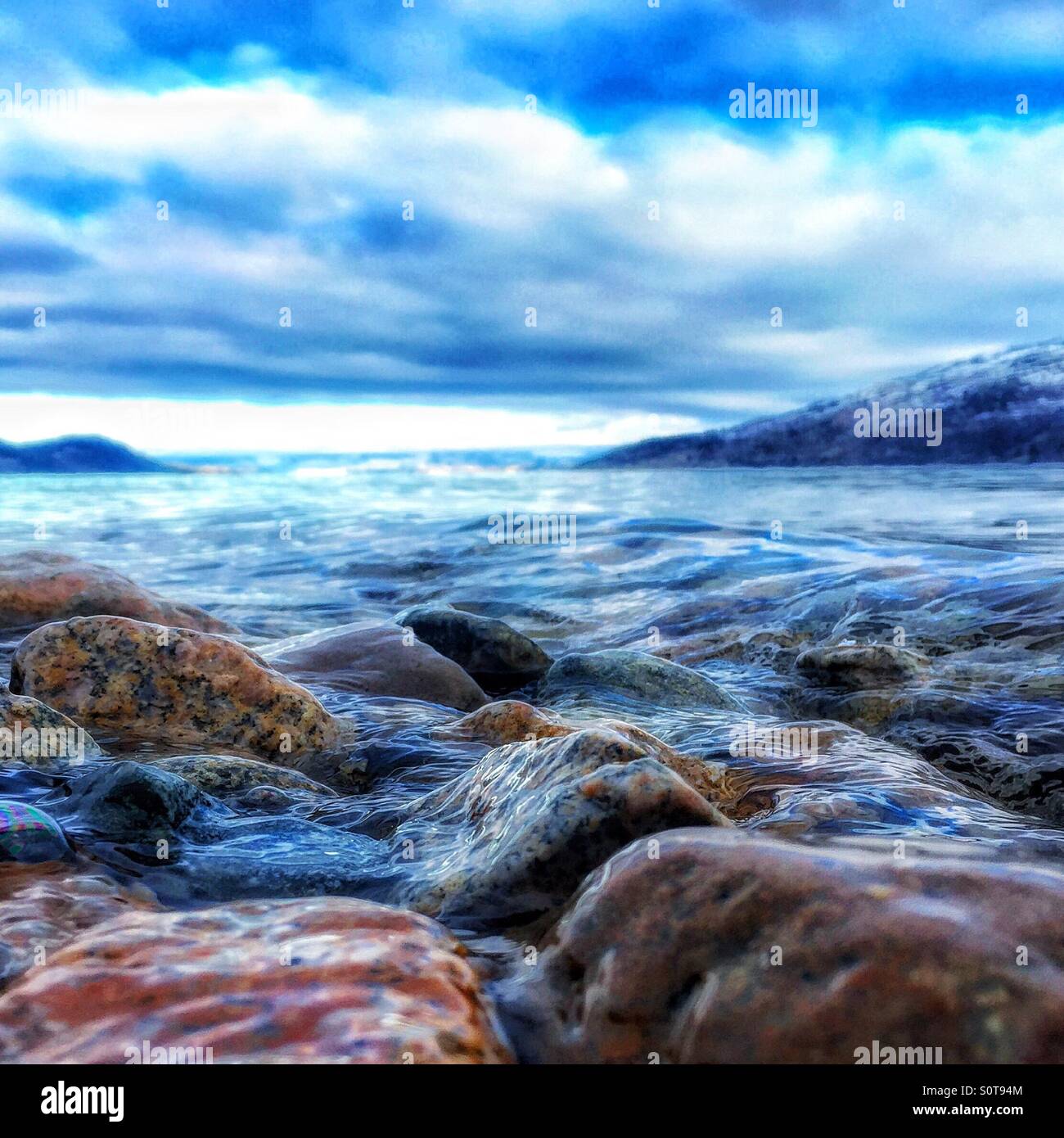 Close up de l'eau à travers de petites roches sur la plage par une journée d'hiver. Montagnes couvertes de neige et ciel nuageux spectaculaires dans l'arrière-plan. - Image de stock capturée avec un smartphone