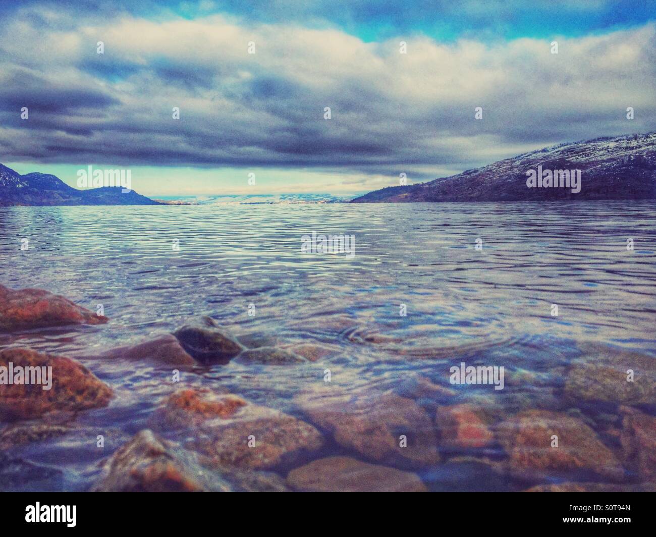 Vue sur le lac Okanagan entouré de montagnes couvertes de neige sous un ciel nuageux ciel d'hiver. Pierres sous l'eau propre au premier plan sont visibles. Banque D'Images