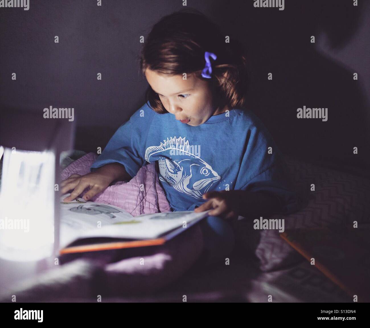 Petite fille lire un livre au lit à la lumière de la lampe Banque D'Images