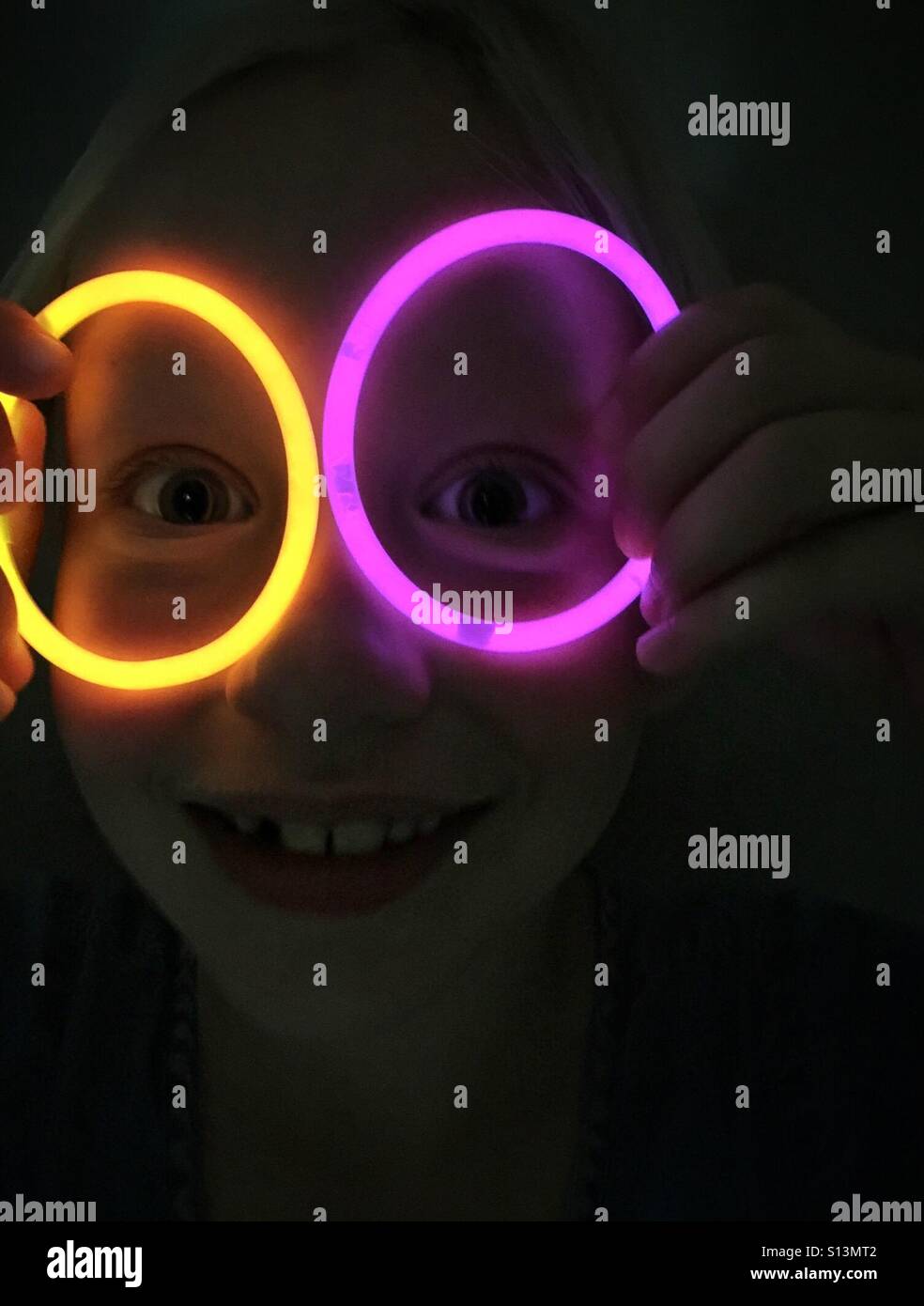 Un enfant ressemble à un clown avec des bâtons lumineux autour des yeux dans une pièce sombre. Banque D'Images