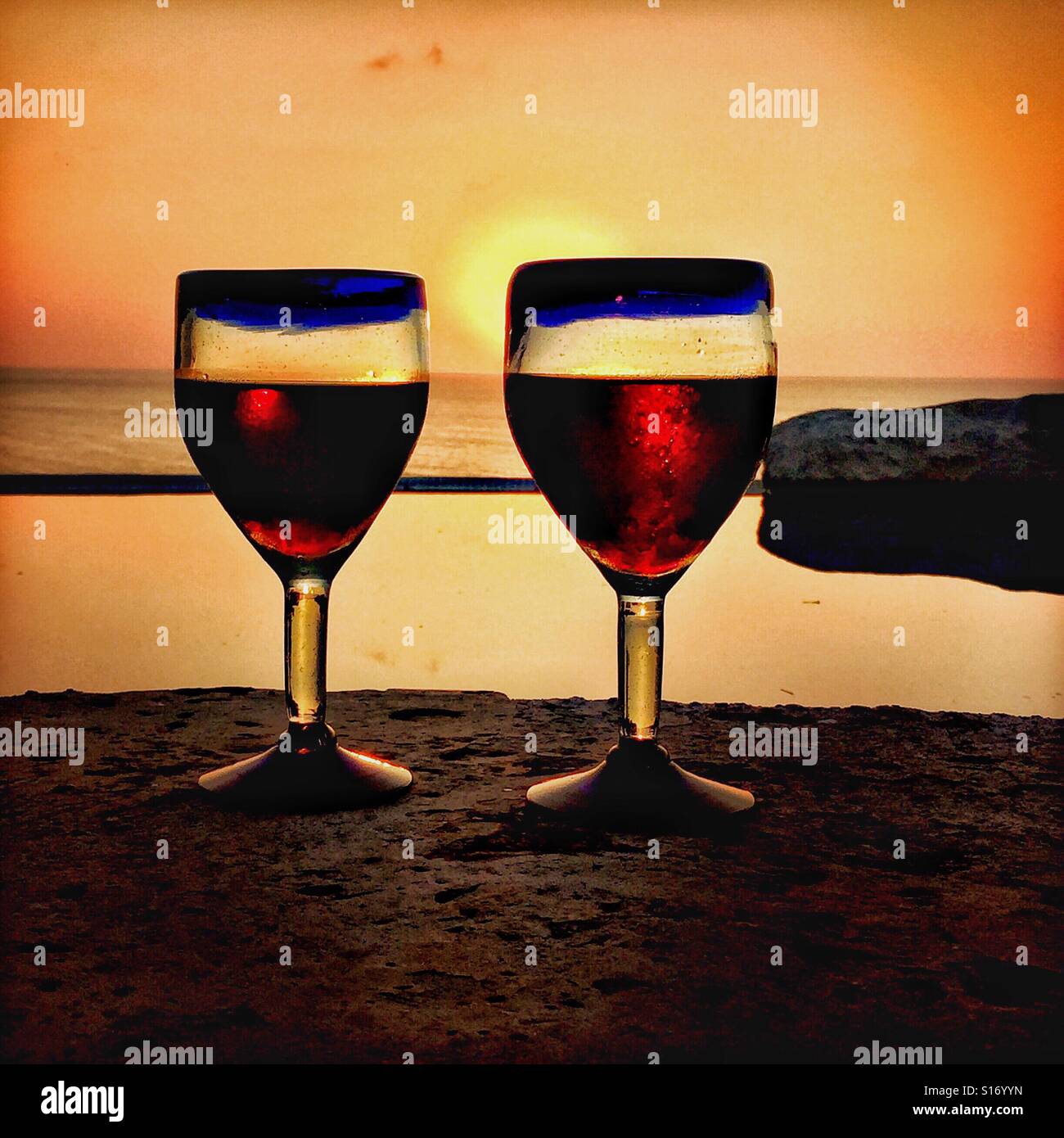 Soufflé à la mexicaine deux paire de lunettes bleu rempli de vin rouge situé en face d'une piscine à débordement avec le soleil sur l'océan Pacifique dans l'arrière-plan dans le Nayarit, Mexique. Banque D'Images