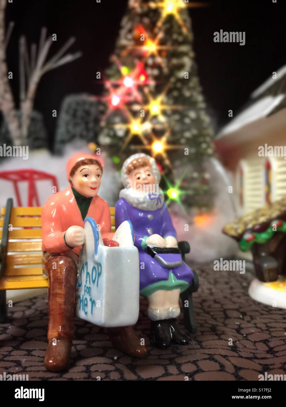 Une figure d'un homme et une femme assis sur un banc à l'extérieur avec un sac d'achats de vacances devant un arbre de Noël décoré de couleurs vives - Image de stock capturée avec un smartphone