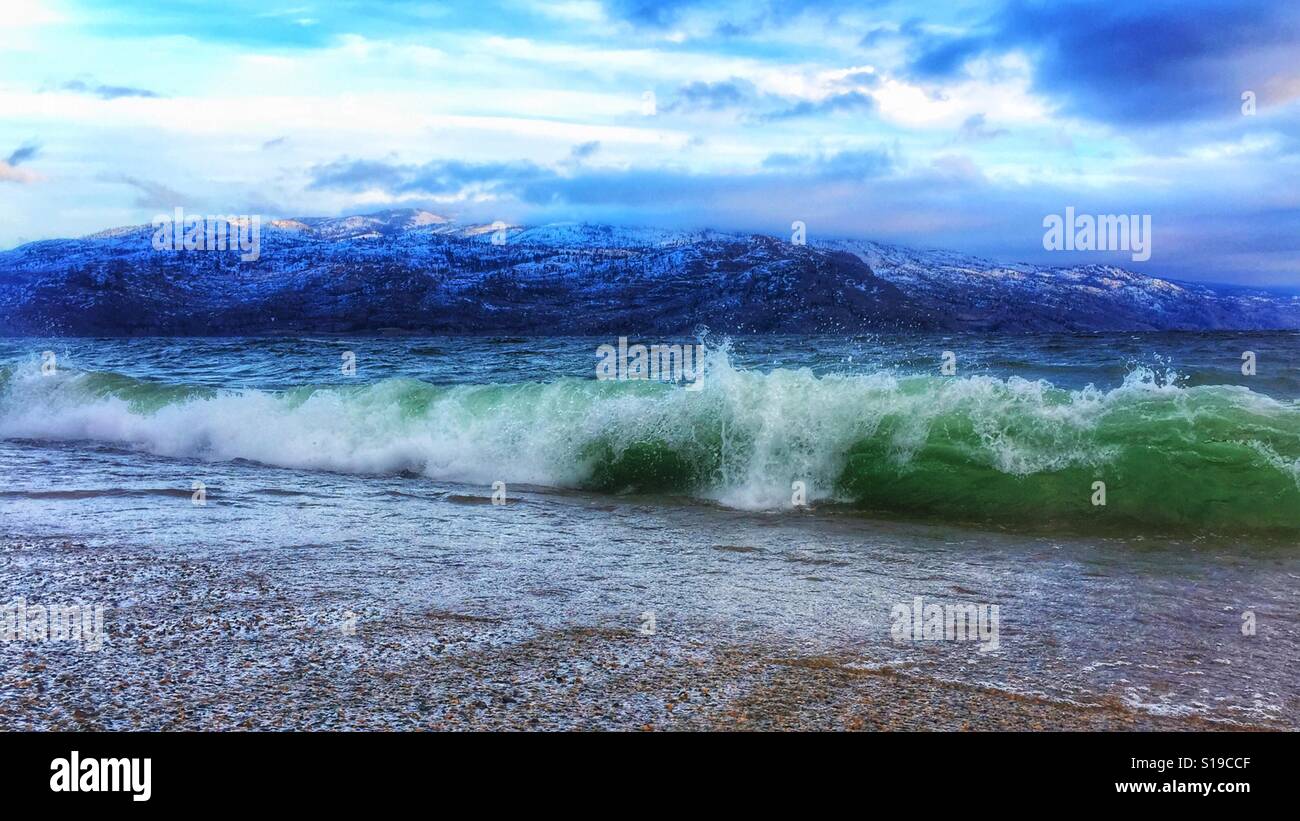 Vue en angle bas des vagues qui s'écrasent sur une plage par une froide journée d'hiver avec des montagnes couvertes de neige au loin - Image de stock capturée avec un smartphone