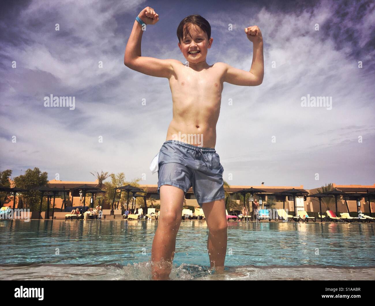 Un garçon de 11 ans fléchit ses muscles après l'escalade d'une piscine extérieure froide pendant vos vacances à Marrakech, Maroc. Banque D'Images