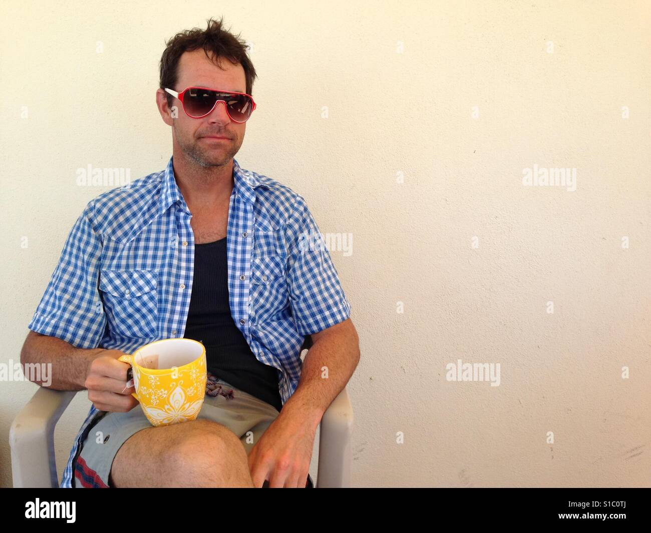 Un homme est assis sur une chaise avec un mug jaune reposant sur ses genoux. Il est en train de regarder la caméra mais portant des lunettes de soleil. Ses cheveux sont ébouriffés et il n'a pas rasé. Banque D'Images