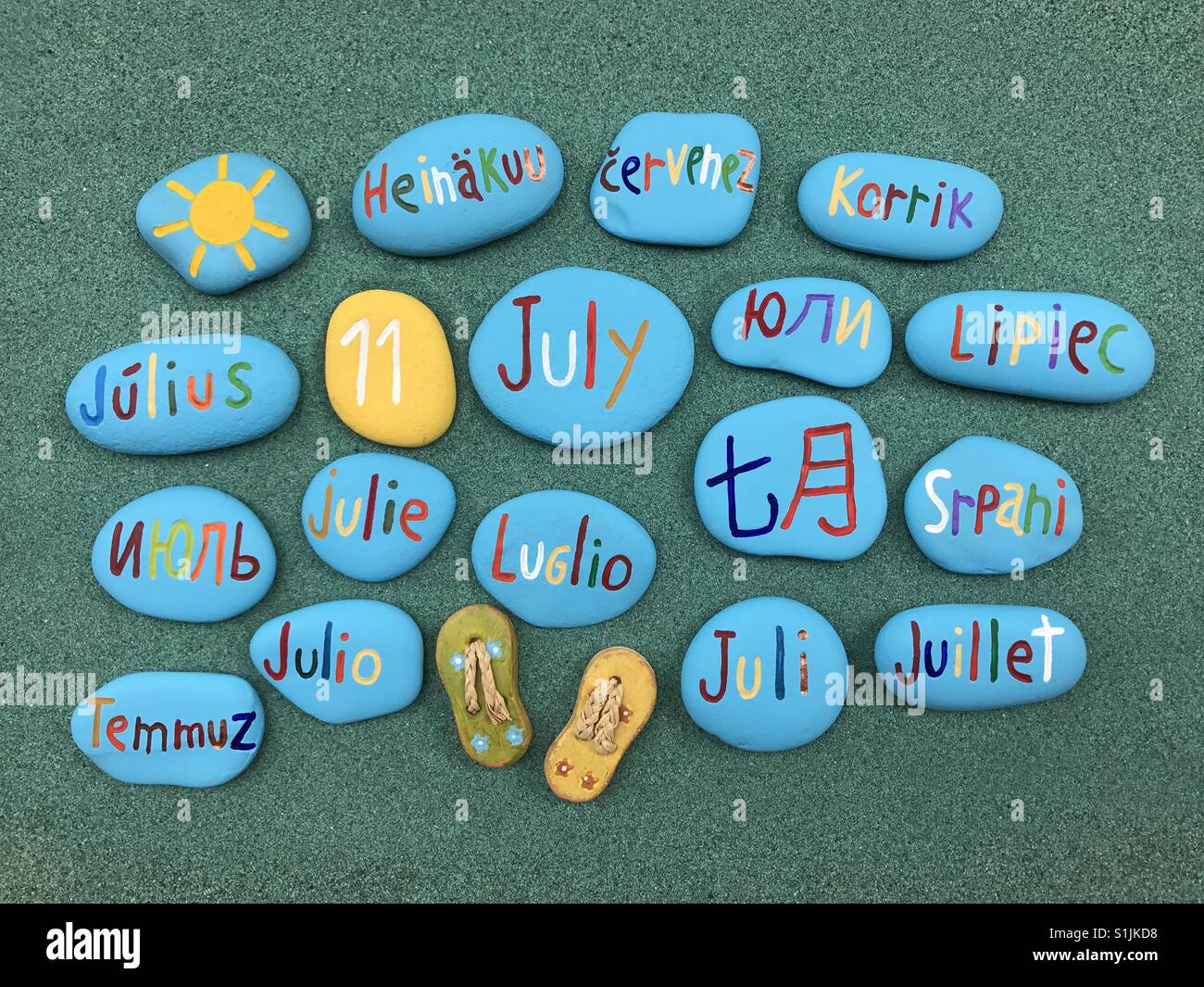 11 juillet, date du calendrier en plusieurs langues sur les pierres de couleur sur le sable vert - Image de stock capturée avec un smartphone
