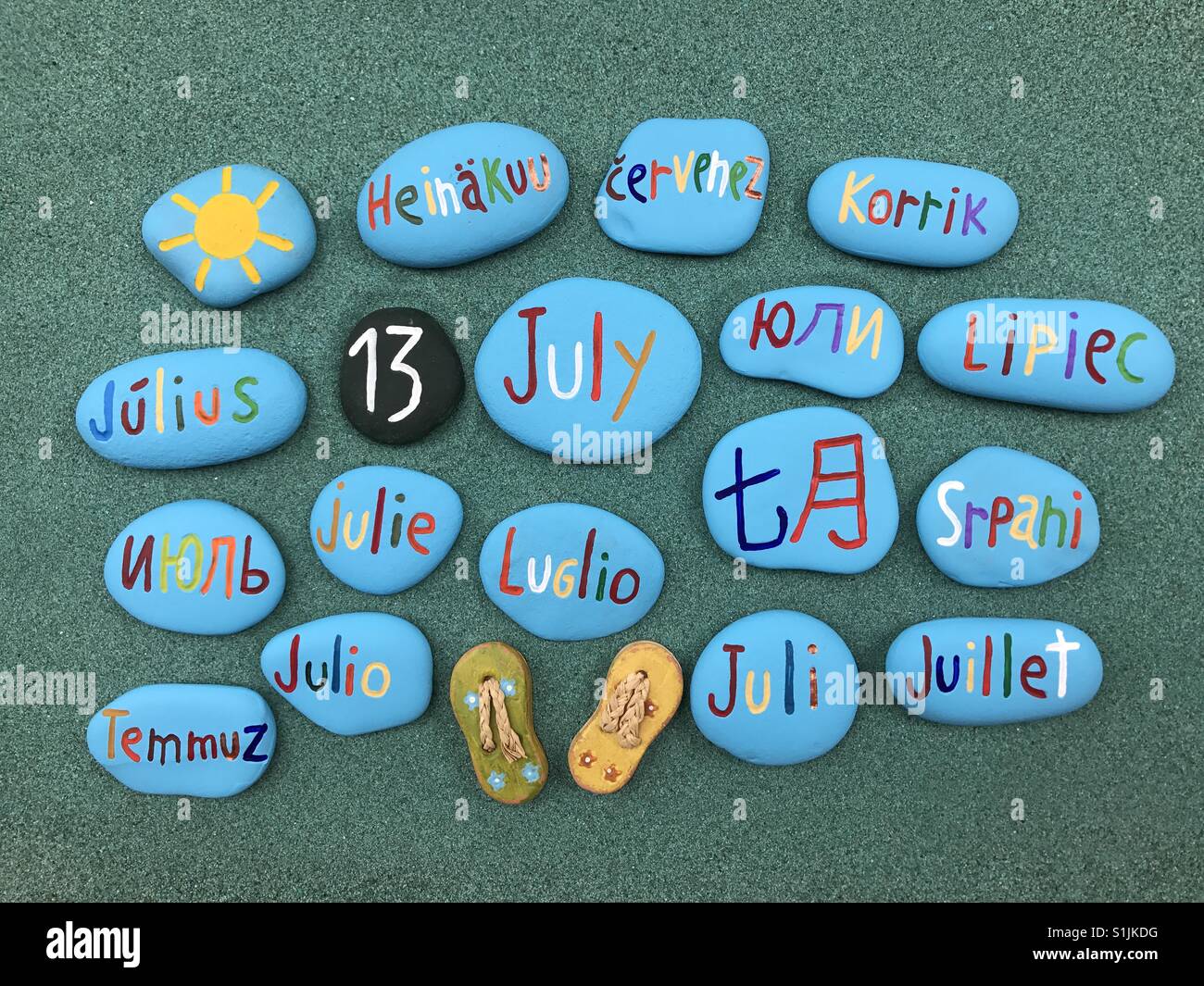 13 juillet, date du calendrier en plusieurs langues sur les pierres de couleur sur le sable vert - Image de stock capturée avec un smartphone