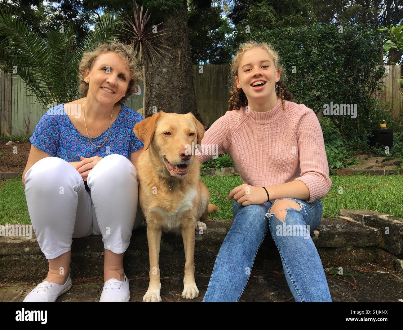 Mère et fille adolescente australienne assis et souriant avec un chien dans un jardin d'accueil - Image de stock capturée avec un smartphone