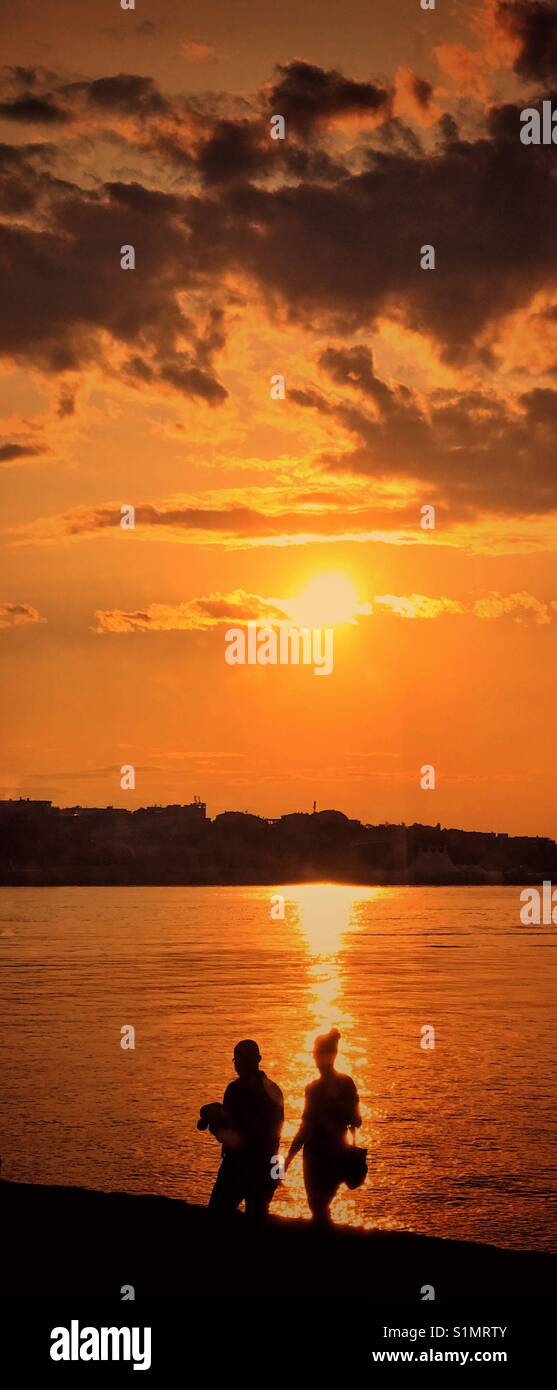 Les silhouettes de deux personnes pendant qu'elles marchent le long d'un mur du port au coucher du soleil. Une image avec des usages multiples. Crédits photos - © COLIN HOSKINS. - Image de stock capturée avec un smartphone