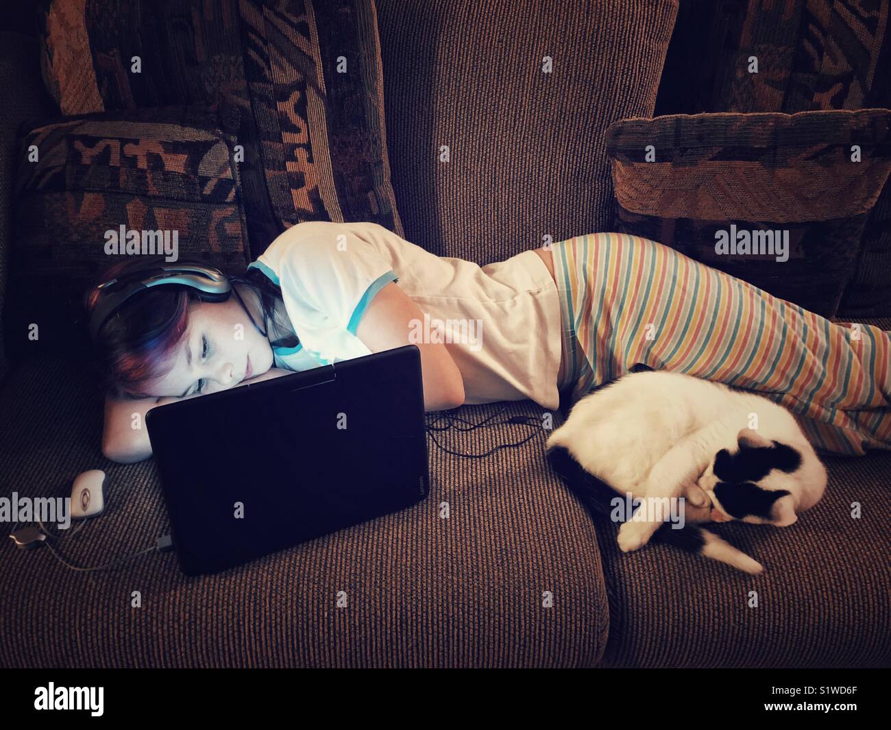 Girl laying on table en utilisant un ordinateur portable avec écouteurs et cat dormir à ses côtés Banque D'Images