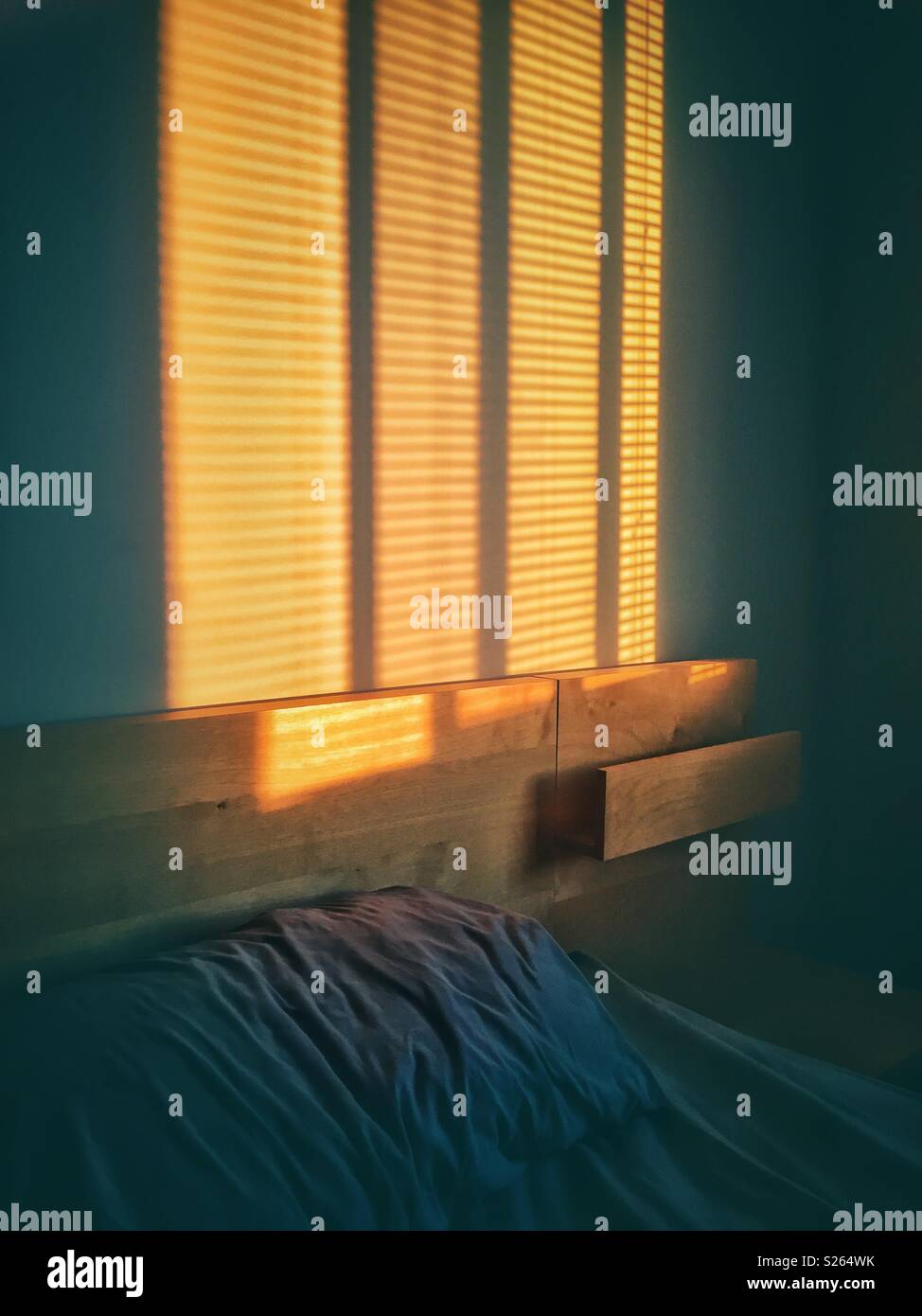 Les patrons de l'orange brillant matin lever du soleil lumière brillant à travers les stores et formant un mur de silhouettes sur chambre et lit défait. Banque D'Images