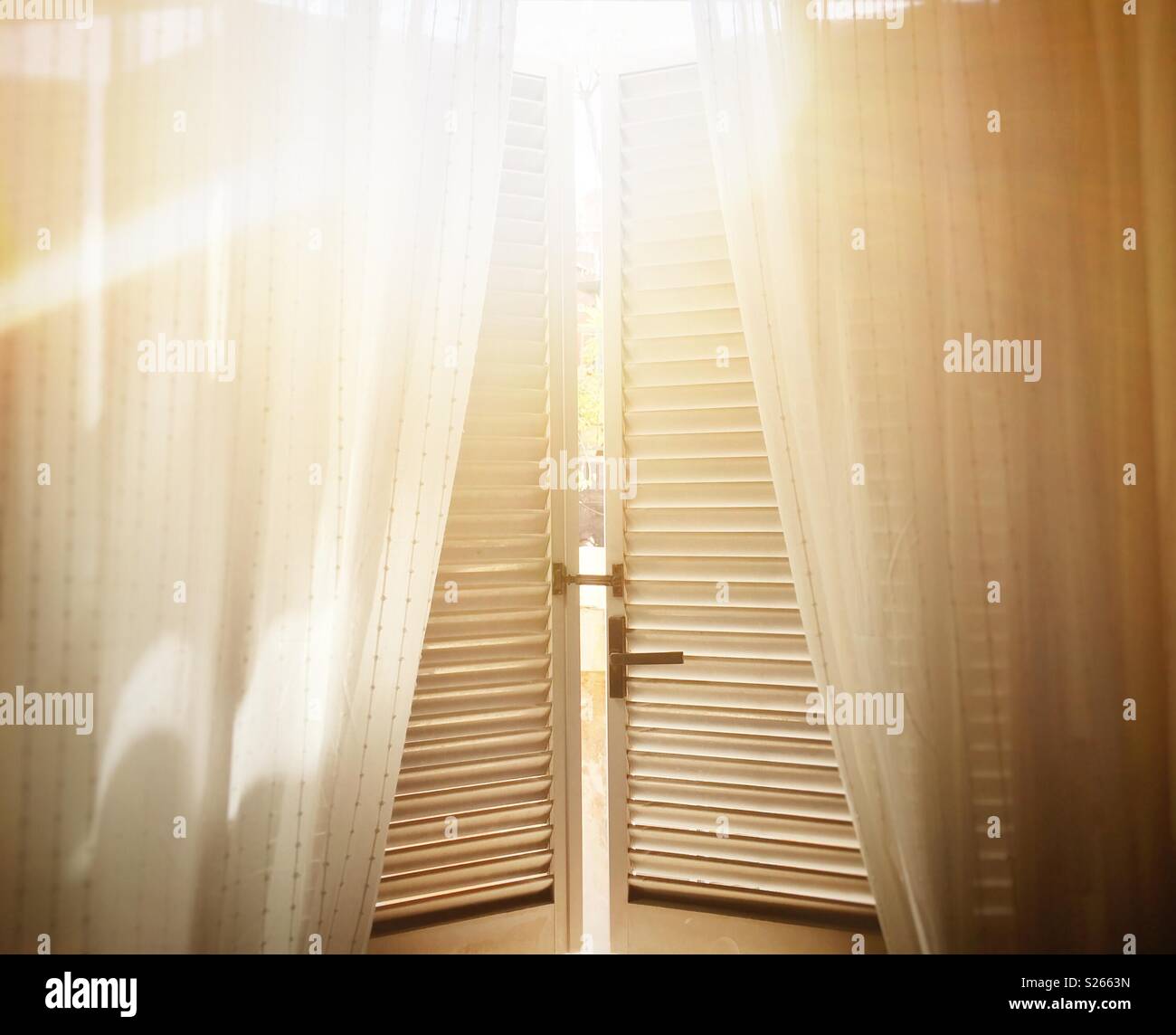 La lumière du soleil filtrée par les volets d'une fenêtre ouverte avec des rideaux en coton blanc Banque D'Images