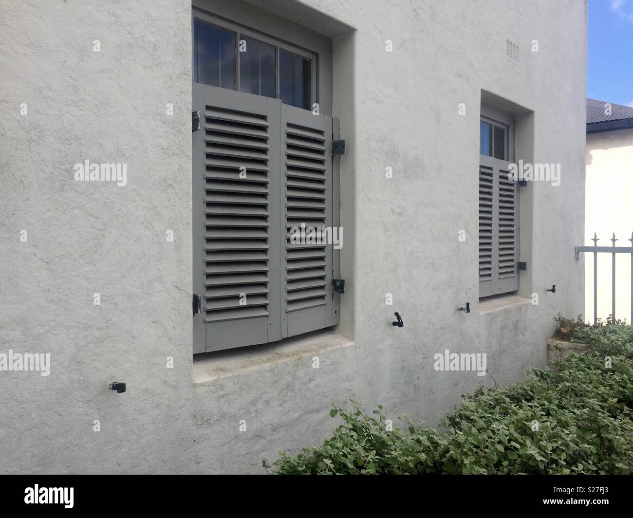 Vue avant de la maison en brique, gris pâle, avec deux fenêtres à demi couvert par des volets en bois peint en gris moyen face à la rue dans la banlieue chic et plantes vertes ci-dessous - Image de stock capturée avec un smartphone