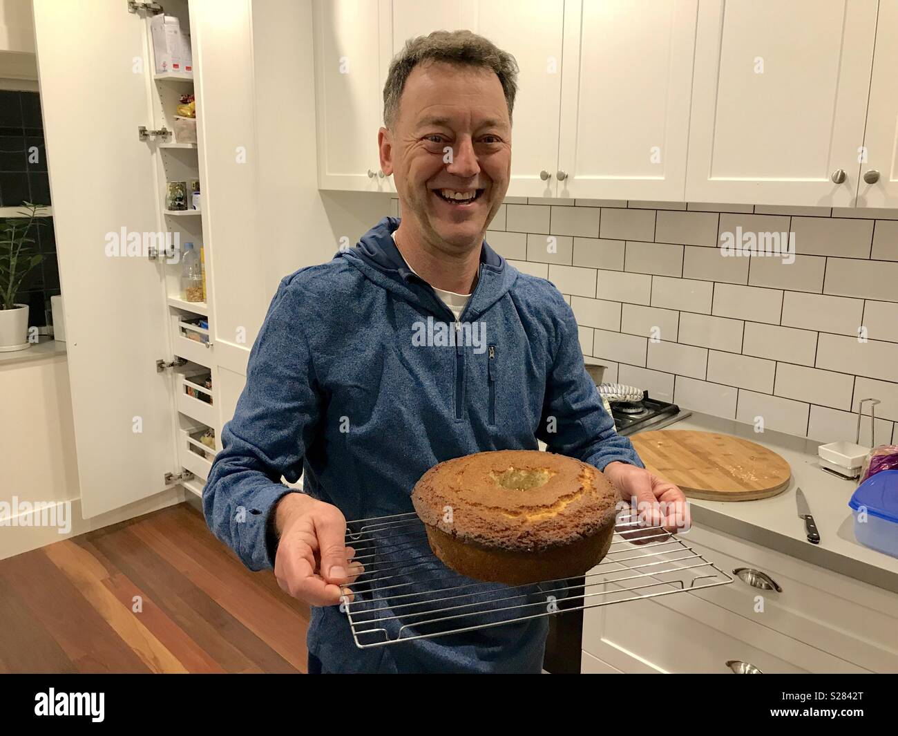 Un homme d'âge moyen blanc debout dans une cuisine moderne tenant un gâteau fraîchement cuit au four - Image de stock capturée avec un smartphone