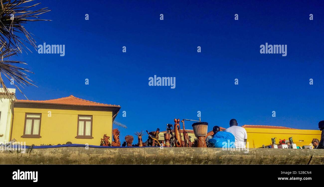 Les vendeurs de rue exposent des objets artisanaux et des marchandises en bois sculptés contre des bâtiments jaunes colorés sous un ciel bleu profond à Espinho, au Portugal. - Image de stock capturée avec un smartphone