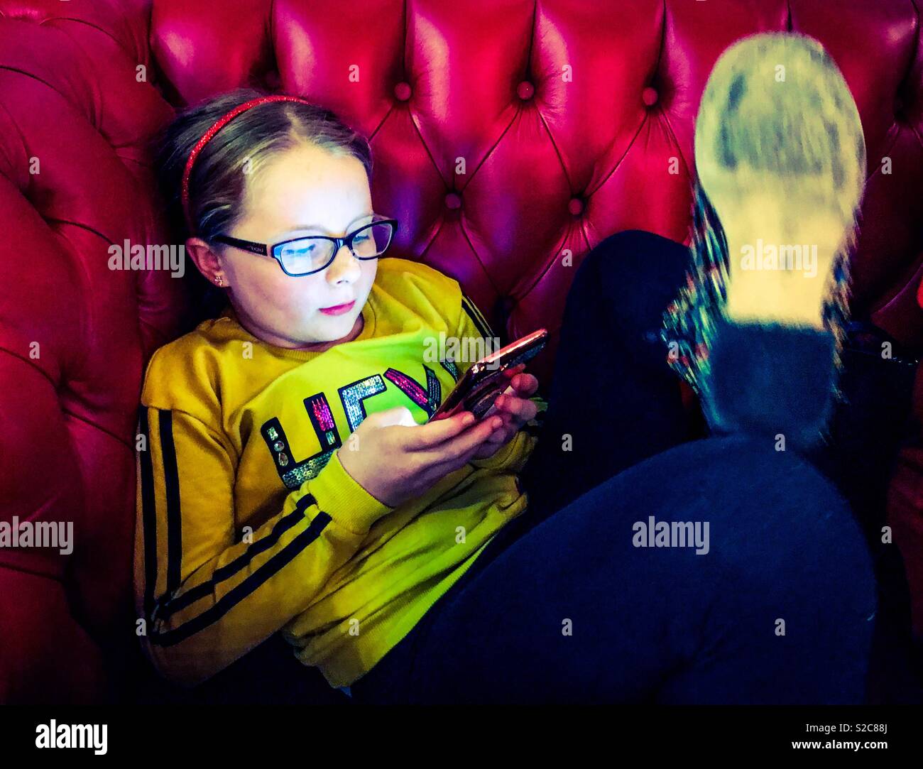 Young Girl playing on smart phone en restant assis sur un canapé en cuir rouge Banque D'Images