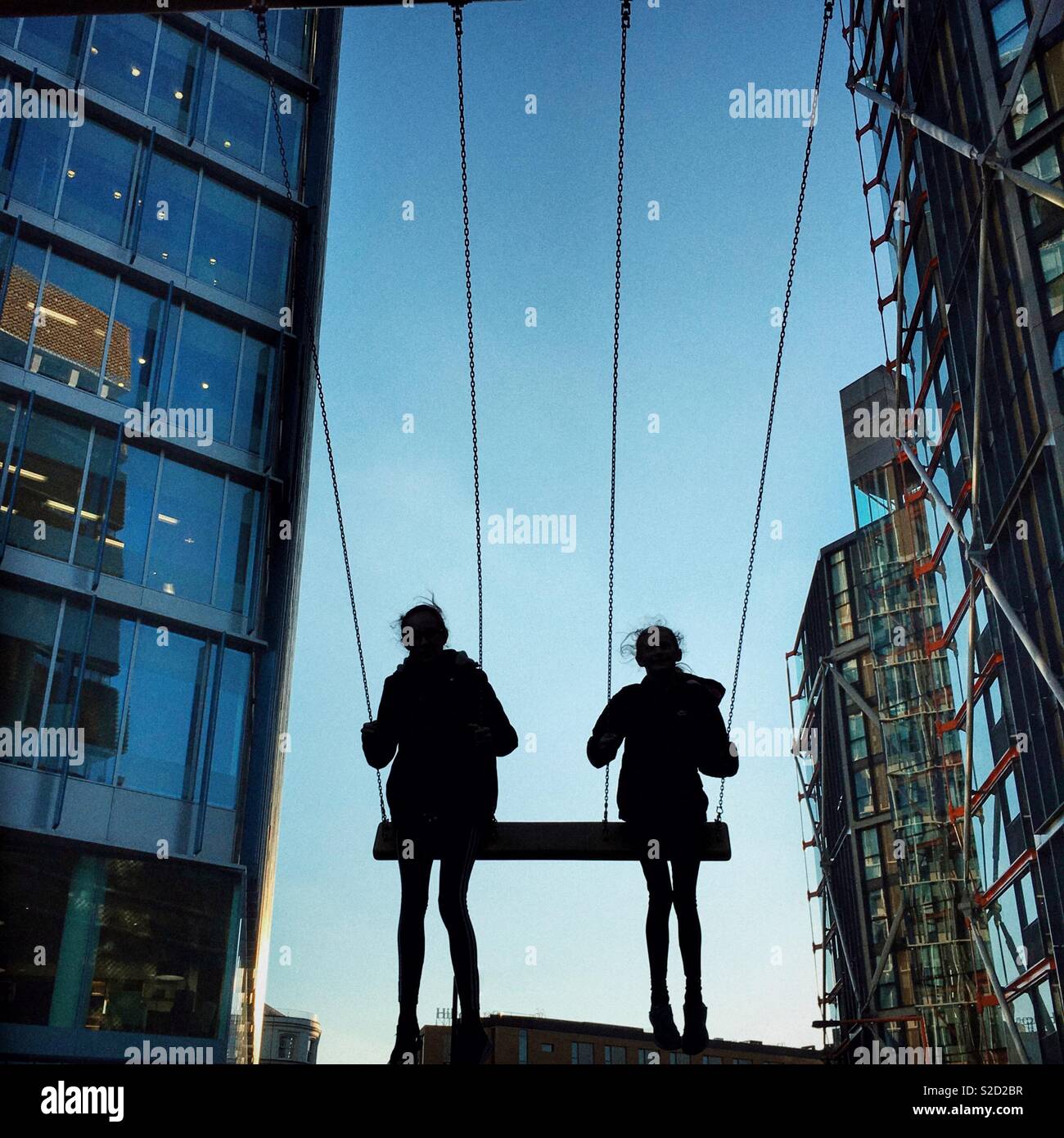 Deux filles sur le grand swing silhouetted against sky avec blocs de bureau moderne d'un côté Banque D'Images
