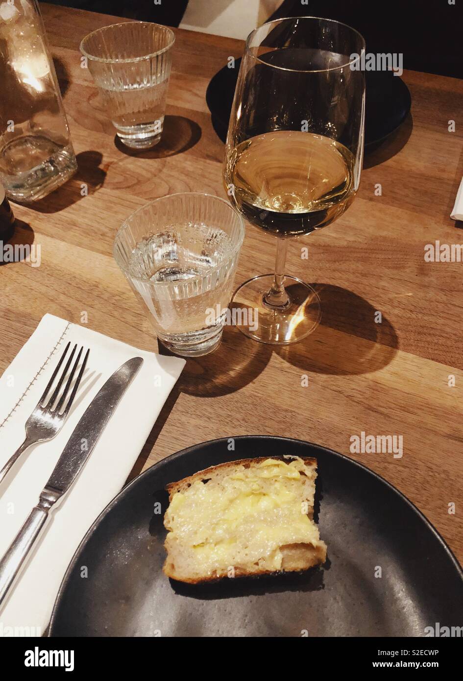 Du pain et du beurre sur la plaque noire mate, le vin blanc et l'eau pétillante avec serviette rustique blanc place setting on wooden table Banque D'Images