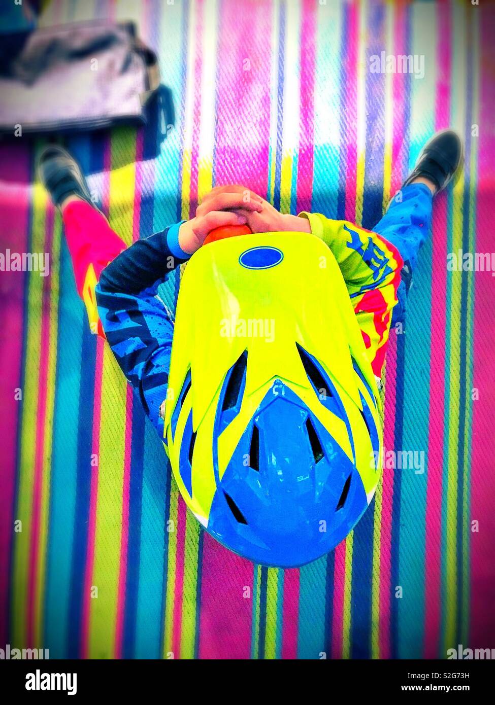 Un enfant dans l'équipement de protection coloré, y compris un casque, est vu de dessus, assis sur un tapis à rayures de couleurs vives. - Image de stock capturée avec un smartphone