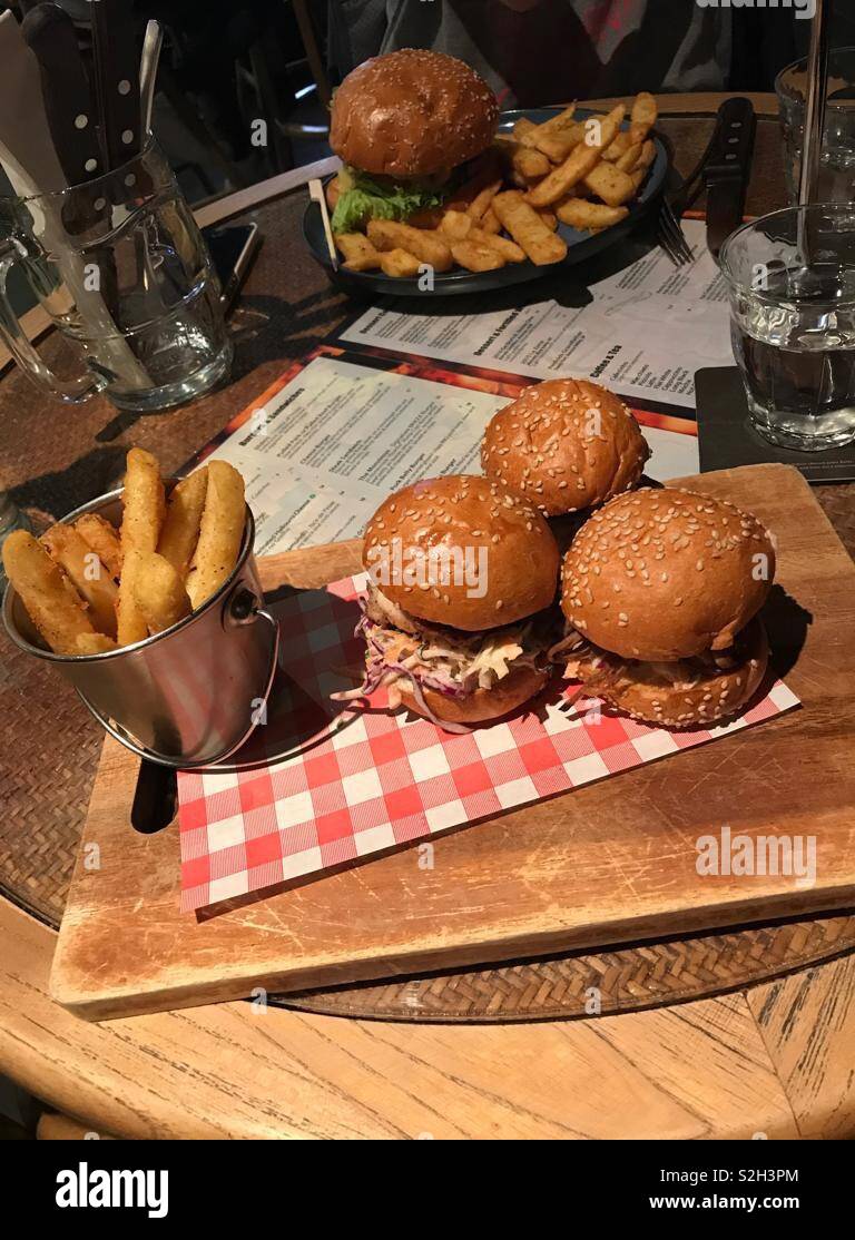 La bonne cuisine moments : De copieux repas rustique d'un trio de hamburgers de brioche avec des hamburgers frites chunky servis sur un plateau en bois avec menu gastro pub derrière et verres d'eau et un autre repas derrière. Banque D'Images