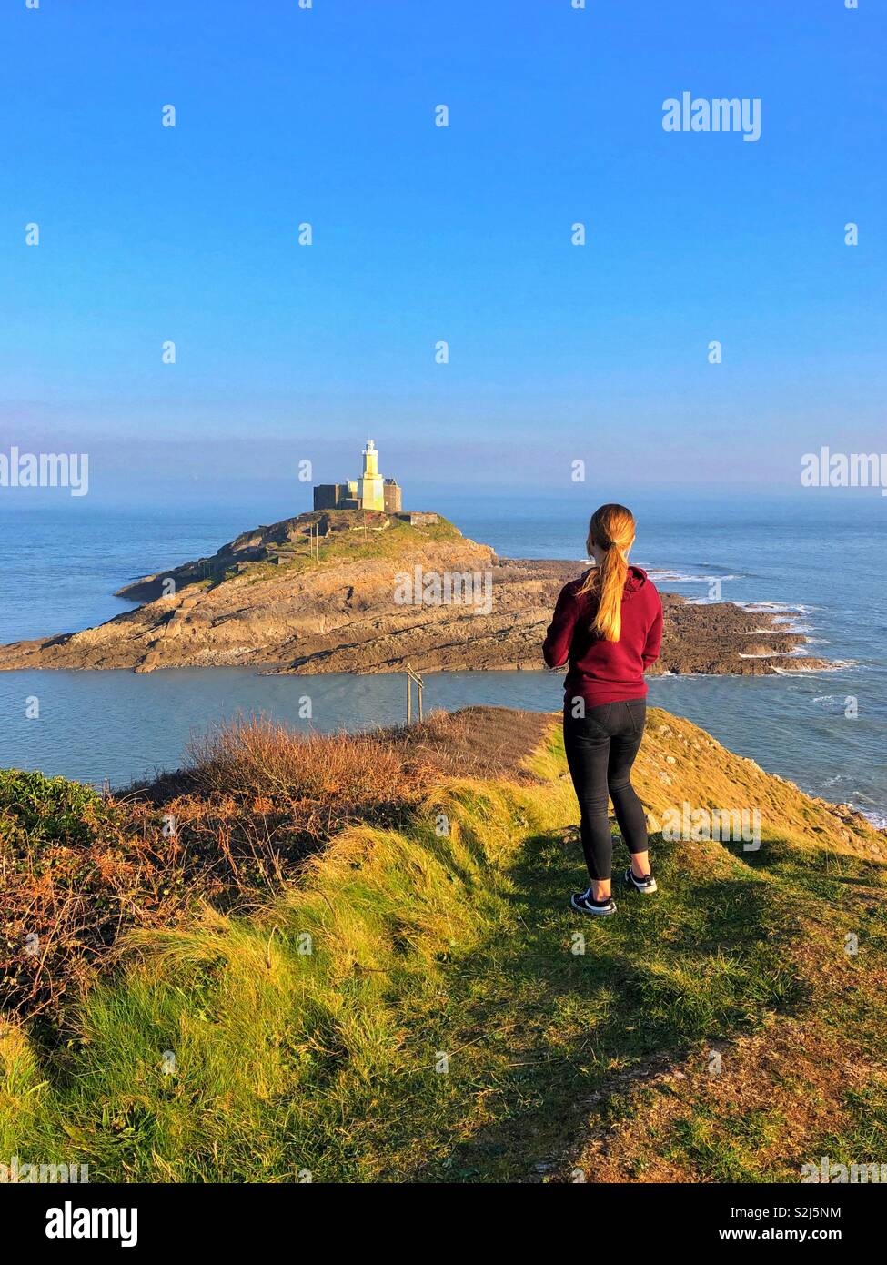Jeune femme à la recherche vers le phare de Mumbles, Mumbles pointe, Swansea, Pays de Galles du Sud-Ouest, février. - Image de stock capturée avec un smartphone