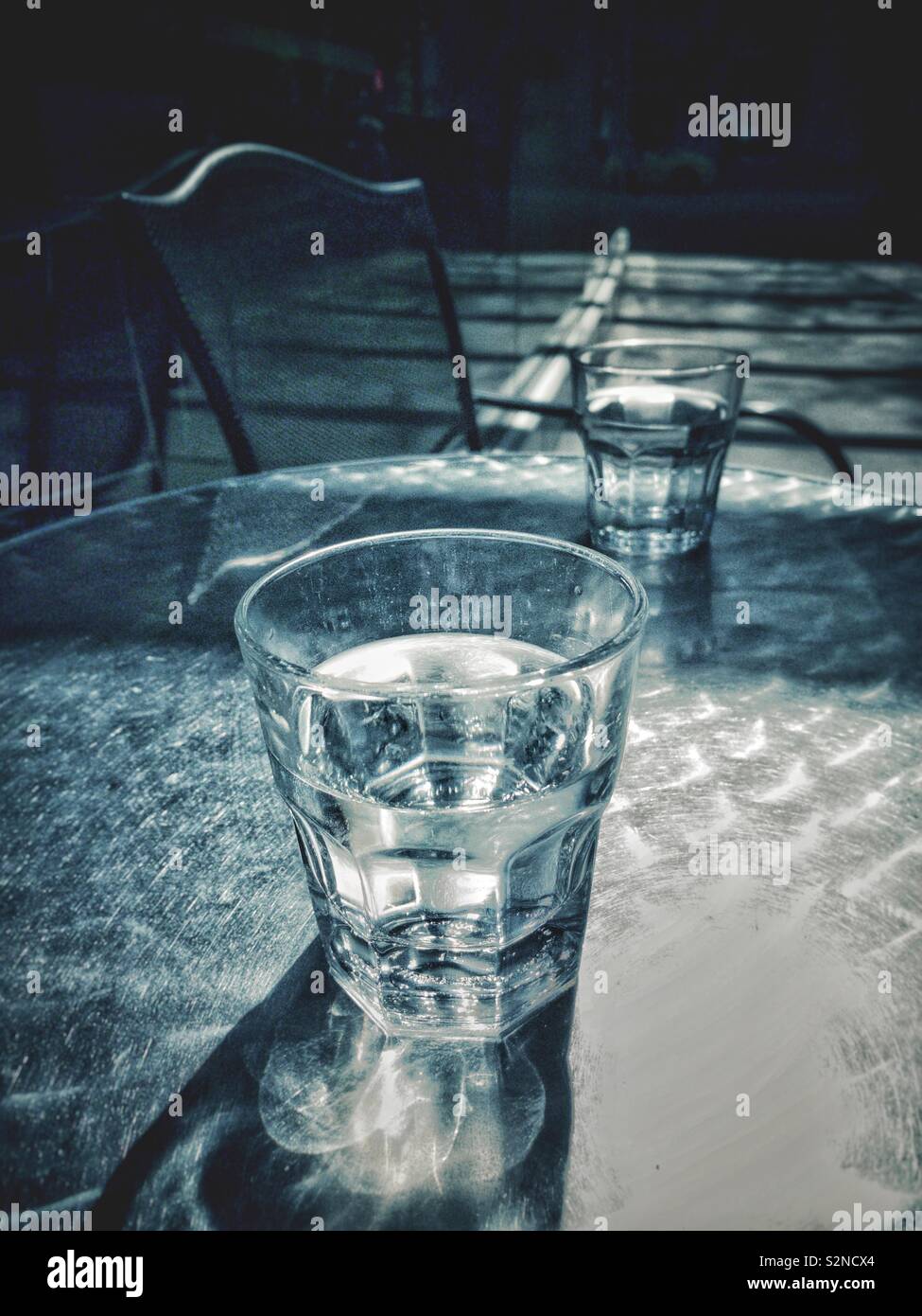 Deux verres d'eau partiellement plein soleil sur la table de patio en métal avec des motifs de la réfraction et l'ombre et une chaise en arrière-plan. - Image de stock capturée avec un smartphone
