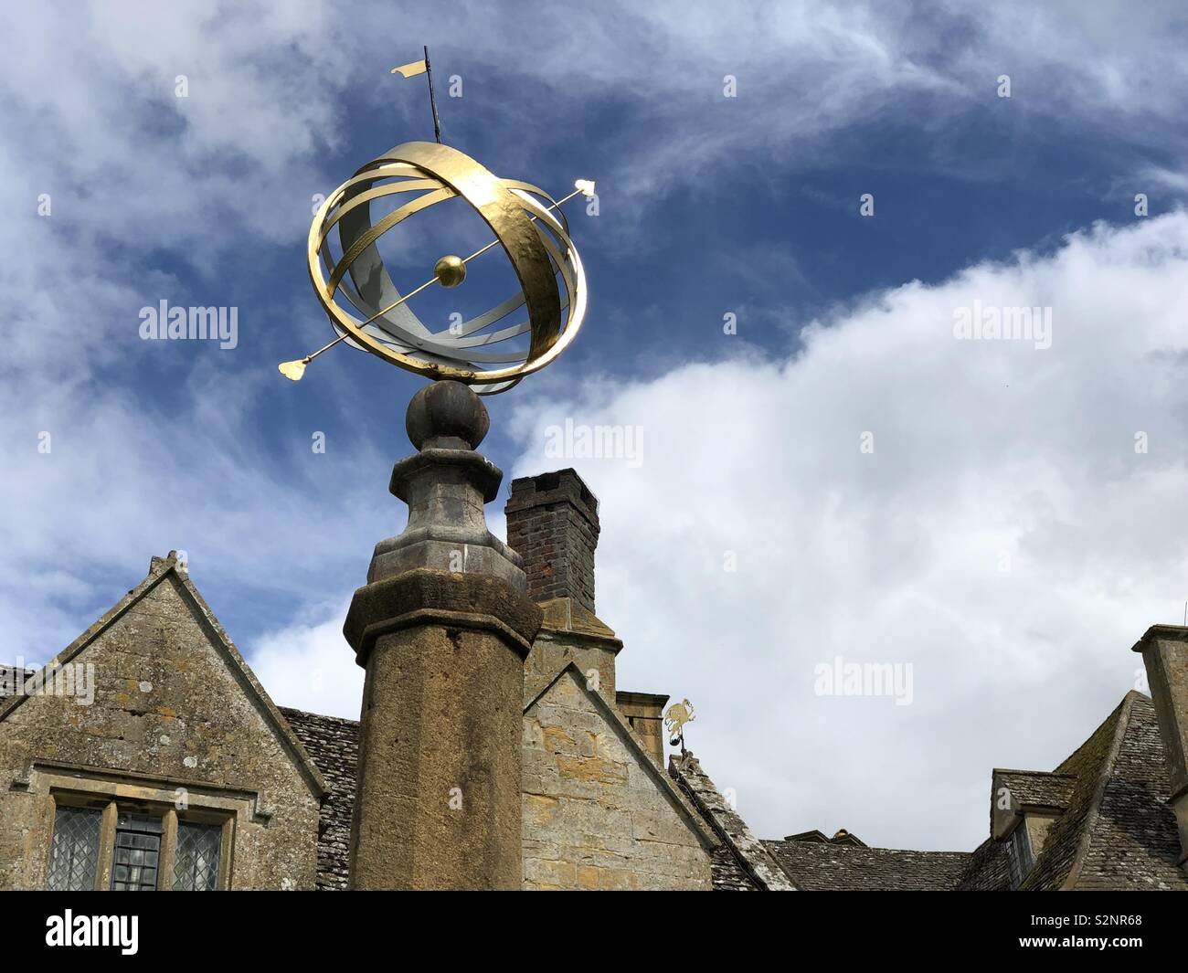 Une sphère armillaire dorée et des lignes de toit Snowshill Manor dans les Cotswolds en contraste avec un ciel bleu vif et des nuages orageux. Banque D'Images