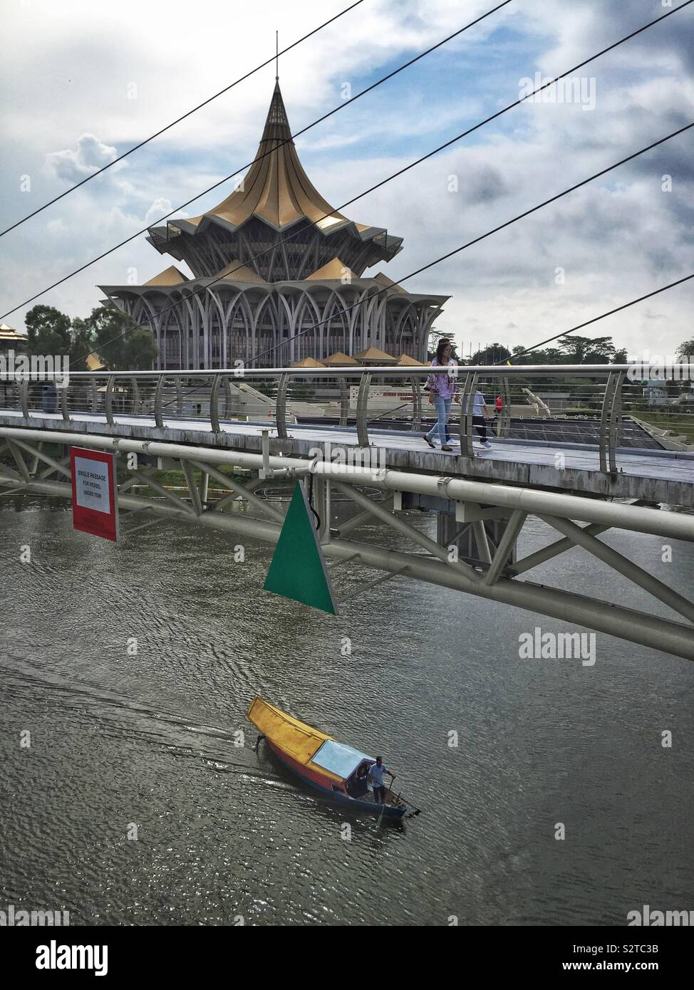 Un sampan malais traditionnel, utilisé pour transporter des personnes à travers la rivière Sarawak, passe sous le pont, Darul Hana avec l'Édifice de l'Assemblée législative de l'État dans l'arrière-plan, Kuching, Malaisie - Image de stock capturée avec un smartphone