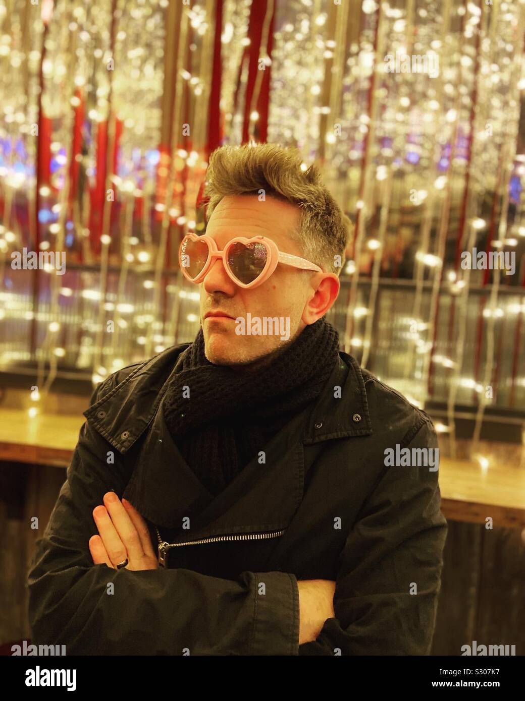 L'homme à la grognon portant des lunettes en forme de coeur rose avec des lumières de Noël en arrière-plan - Image de stock capturée avec un smartphone