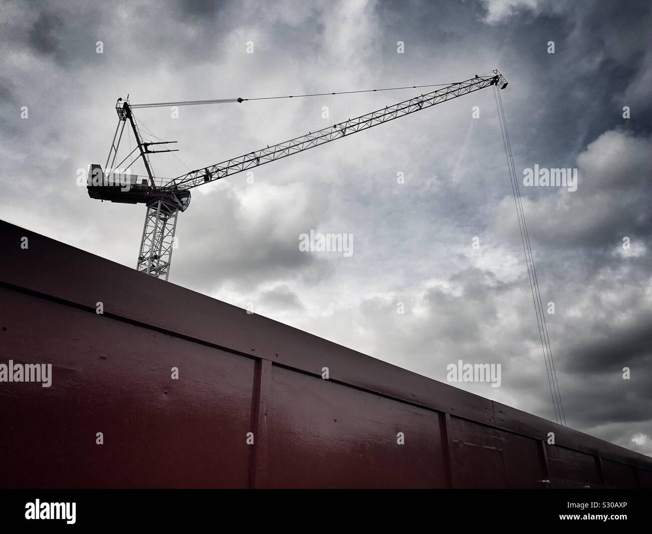 Grue de construction derrière un mur de chantier contre un ciel dramatique. Banque D'Images
