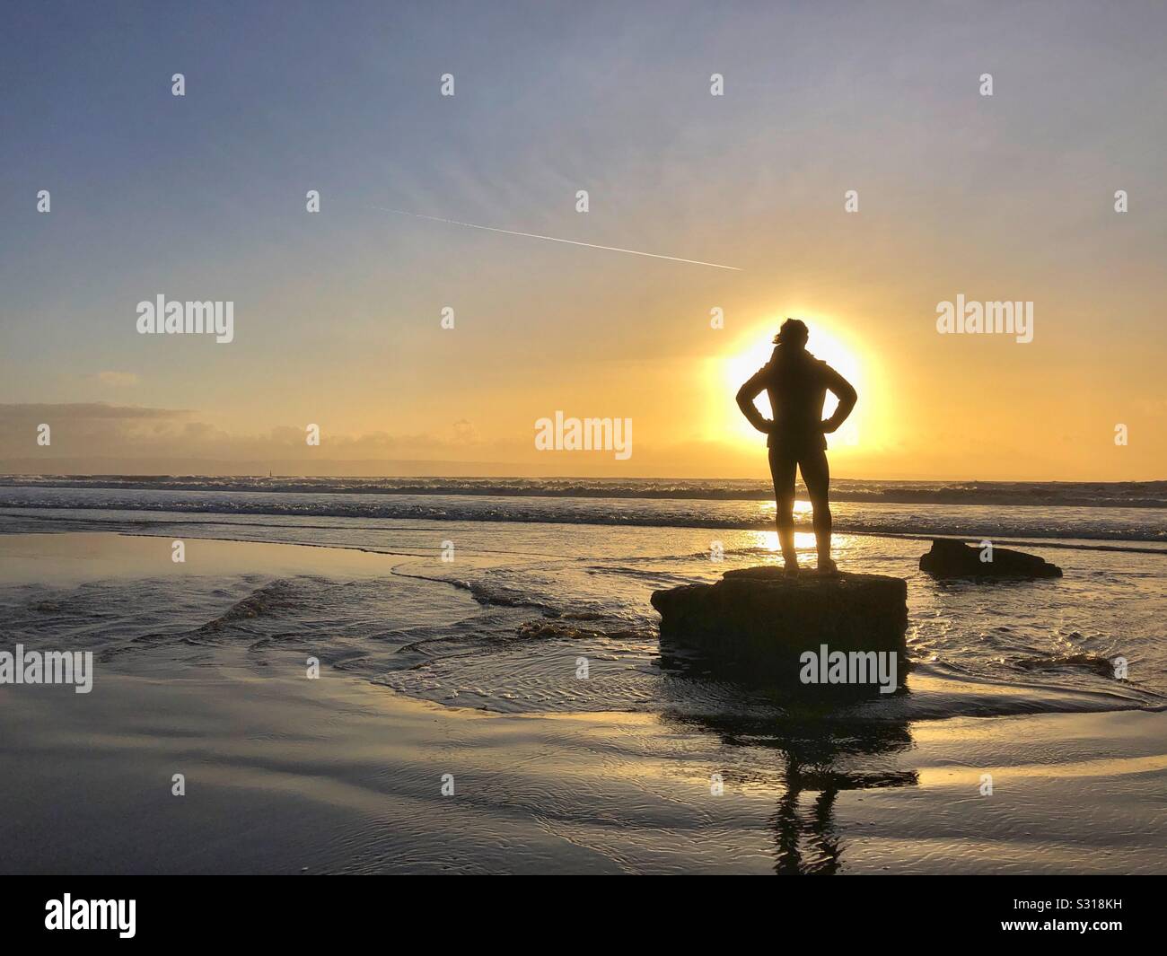 Jeune femme en silhouette debout sur un rocher au bord de la mer au coucher du soleil, dans le sud du Pays de Galles, janvier. Banque D'Images