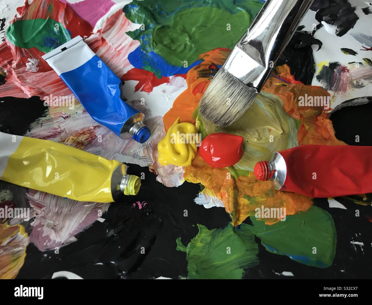 Palette d'artistes avec pinceau mélangeant les couleurs rouge et jaune et les tubes de peinture dispersés autour. Banque D'Images