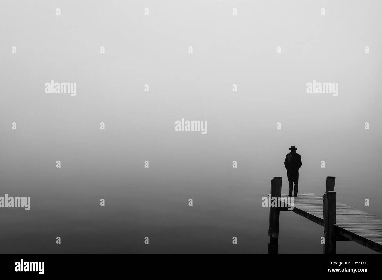 Isolement social : une figure masculine solitaire avec chapeau se tient sur un quai au-dessus d'un lac misty Banque D'Images