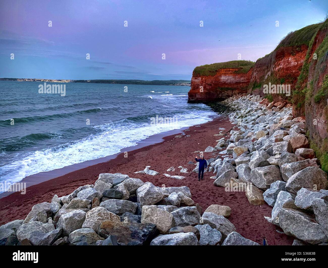 Homme debout sur une plage à marée haute, entouré de falaises rouges, de sable, de grands rochers et d'une mer orageux. - Image de stock capturée avec un smartphone