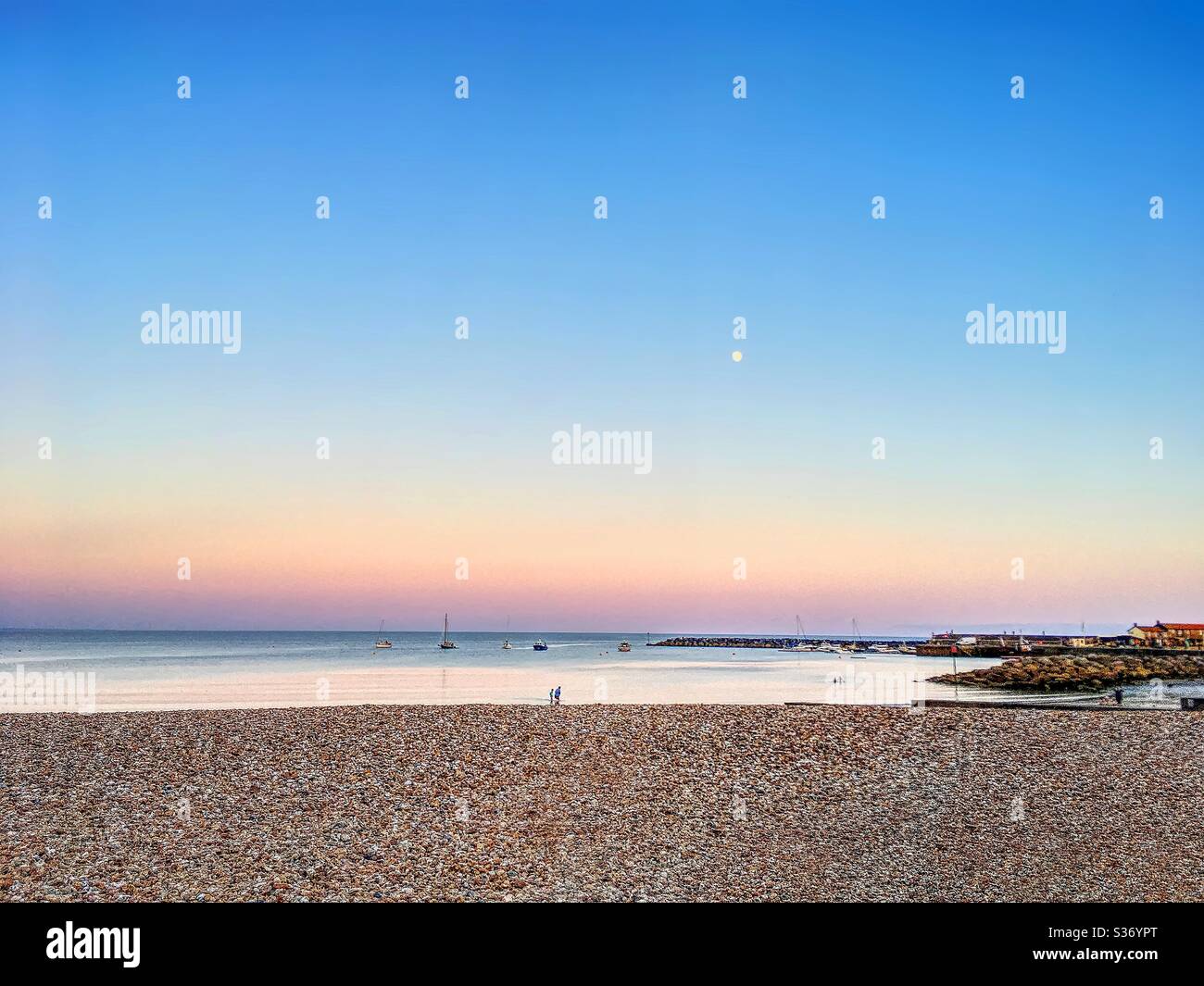 Crépuscule se rassemblant sur une mer calme avec une surface semblable à un miroir. Bleu profond, jaune et violet dans le ciel, et une petite lune. Plage de galets avec de minuscules chiffres. Petit artisanat juste à l'extérieur de la mer. Maisons éloignées. Banque D'Images