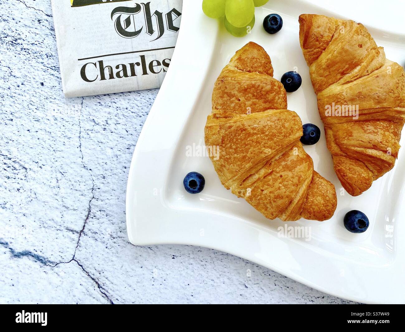 Vue rapprochée des croissants fraîchement cuits sur une assiette blanche aux myrtilles et aux raisins. Petit déjeuner savoureux avec journal du matin sur une table en pierre de granit naturel. - Image de stock capturée avec un smartphone