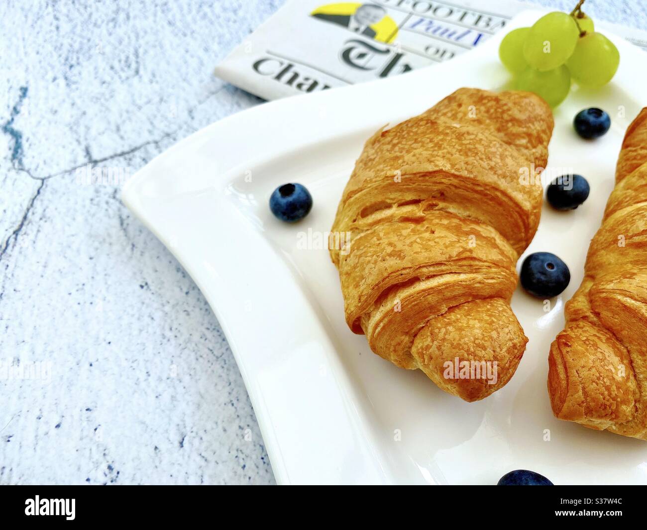 Vue rapprochée des croissants fraîchement cuits sur une assiette blanche aux myrtilles et aux raisins. Petit déjeuner savoureux avec journal du matin sur une table en pierre de granit naturel. - Image de stock capturée avec un smartphone