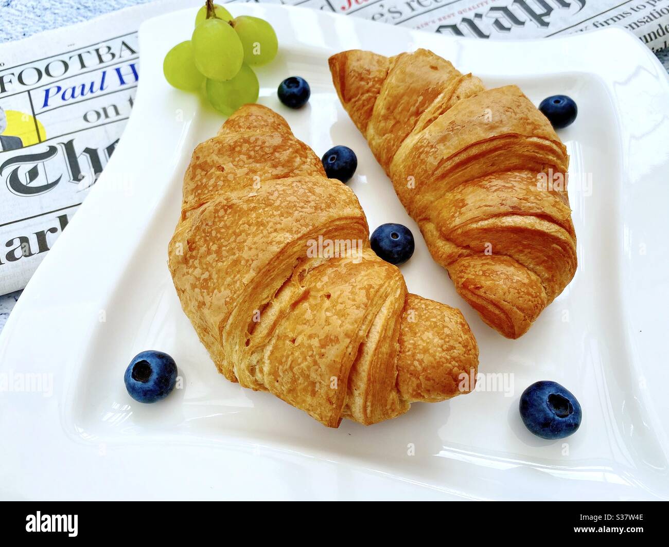 Vue rapprochée des croissants fraîchement cuits sur une assiette blanche aux myrtilles et aux raisins. Petit déjeuner savoureux avec journal du matin sur une table en pierre de granit naturel. Banque D'Images