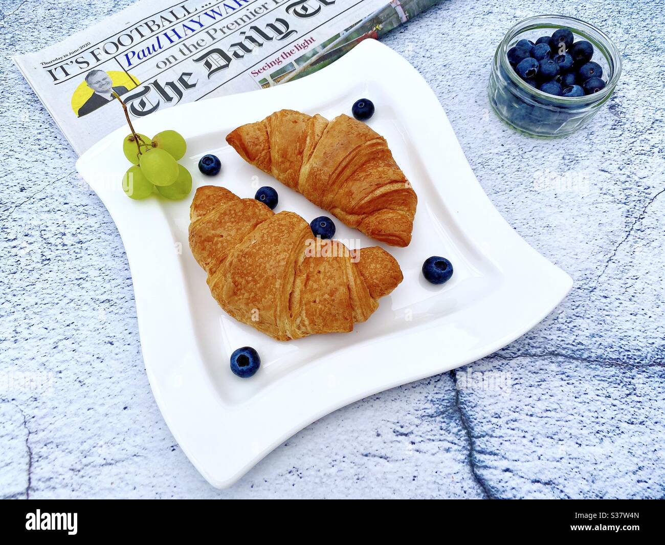 Vue rapprochée des croissants fraîchement cuits sur une assiette blanche aux myrtilles et aux raisins. Petit déjeuner savoureux avec journal du matin sur une table en pierre de granit naturel. Banque D'Images
