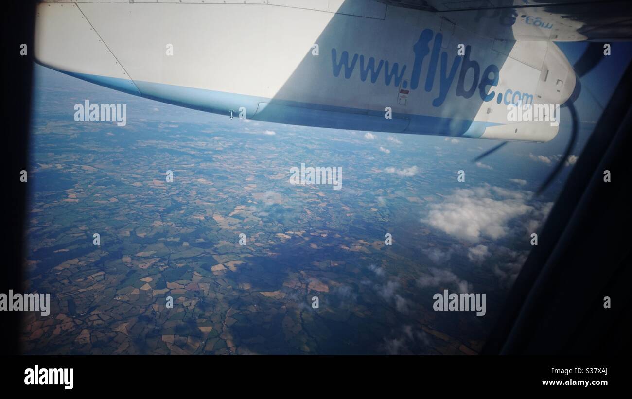 Une photo de l'intérieur d'un avion Flybe, qui regarde par-dessus la terre en contrebas, survolant les villes et les villages, lors d'une belle journée ensoleillée avec quelques nuages. Banque D'Images