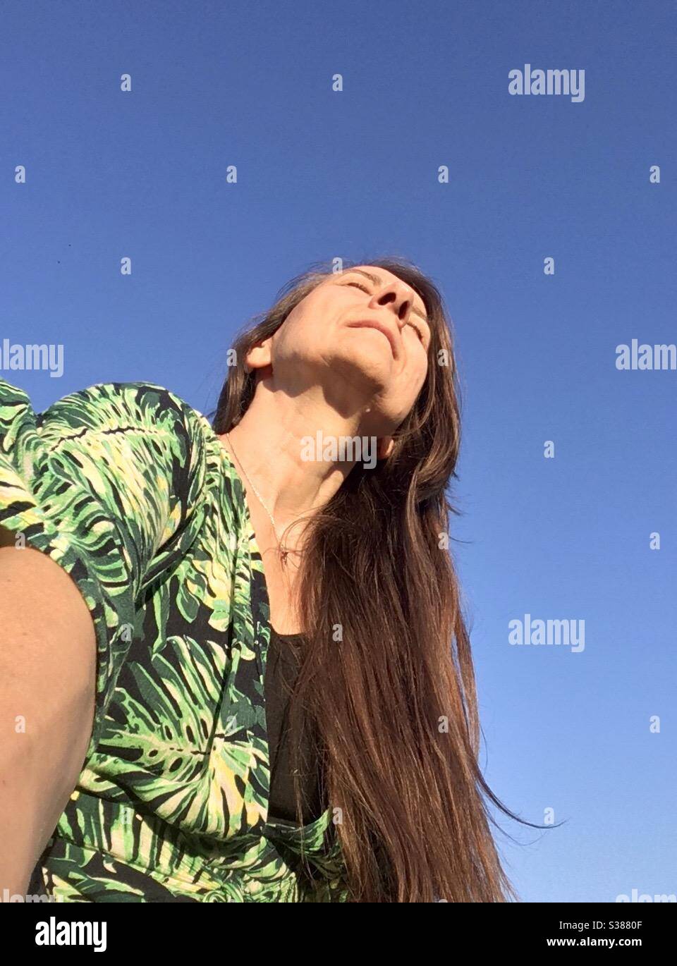 Selfie, femme aux longs cheveux droits dans une robe d'été lumineuse appréciant le soleil brillant sur son visage avec le ciel bleu parfait comme arrière-plan, modèle disponible Banque D'Images