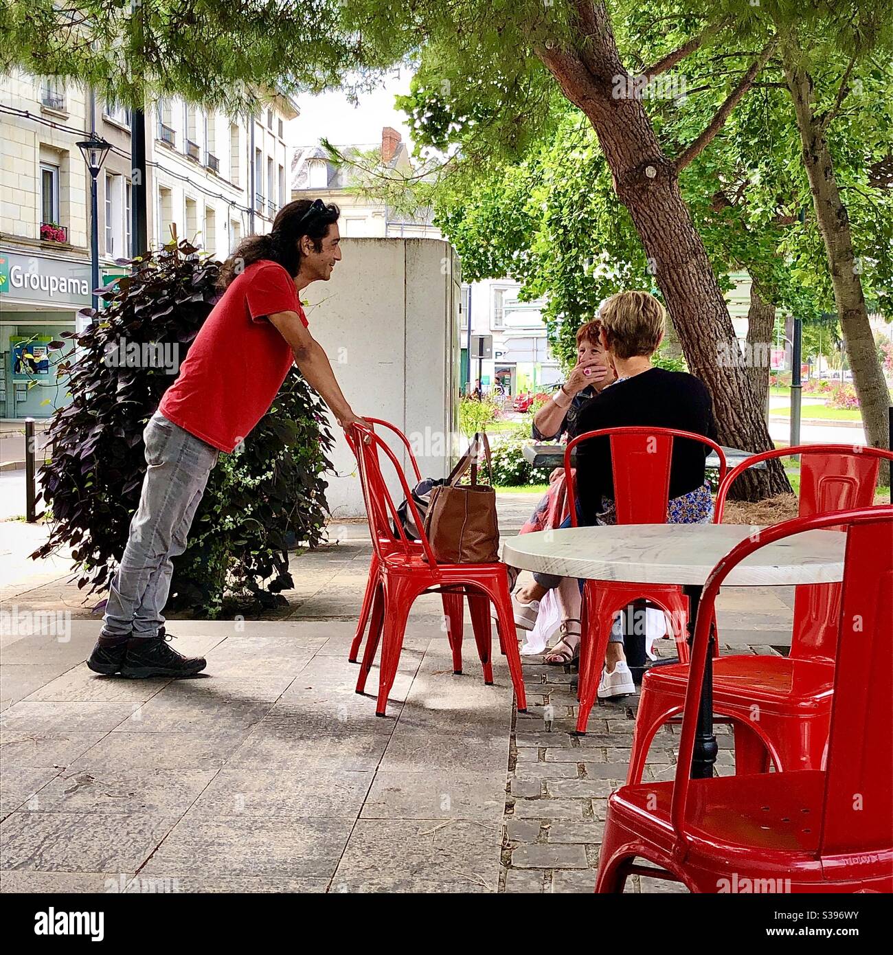 Homme penché à angle impair contre une chaise parlant aux femmes sur une terrasse de café - France. - Image de stock capturée avec un smartphone