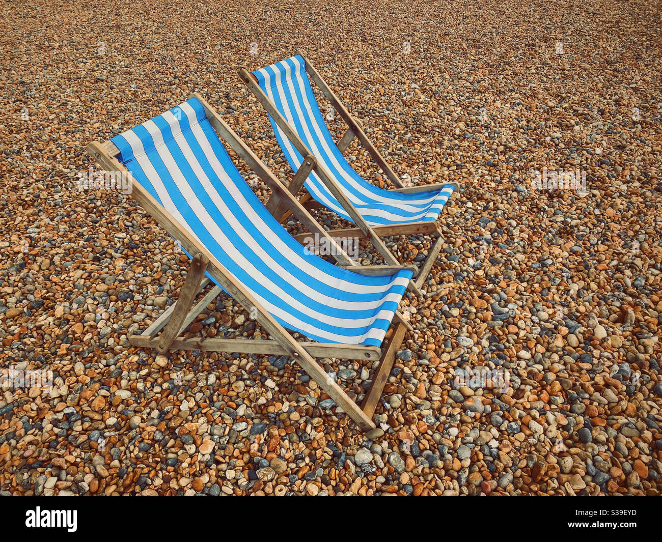 Deux chaises longues en bois à rayures bleues et blanches - toutes seules et entourées de galets de plage. Où sont tous les gens? Photo ©️ COLIN HOSKINS. Banque D'Images