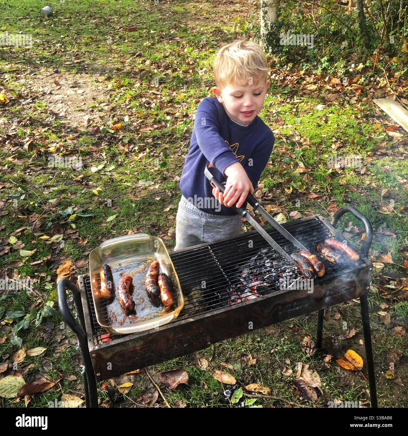 Un garçon de quatre ans cuisant des saucisses sur un barbecue. - Image de stock capturée avec un smartphone