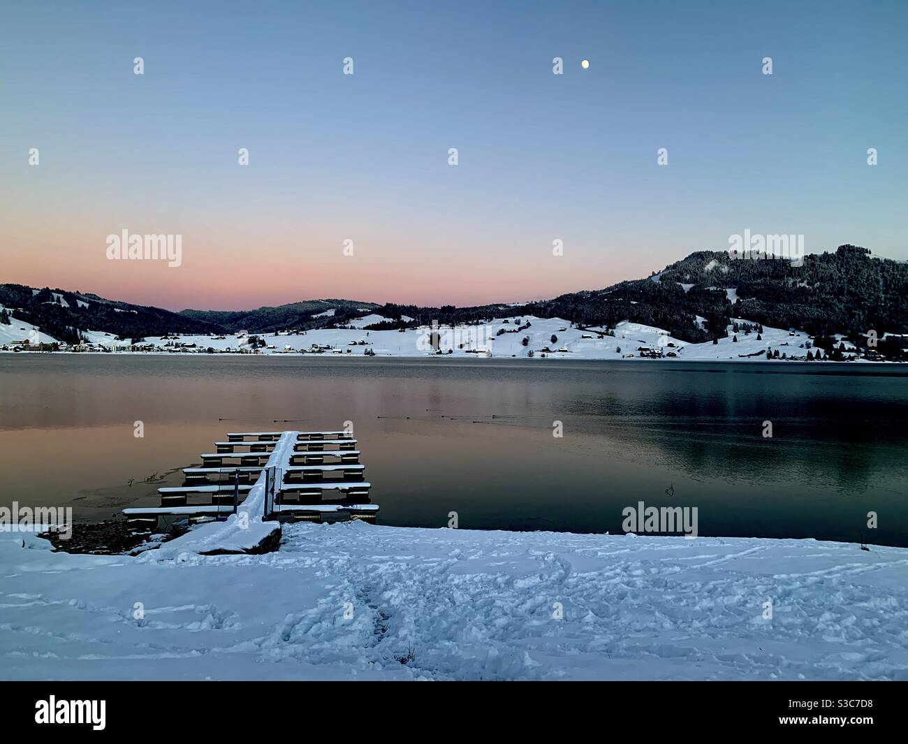 Coucher de soleil sur le lac Sihl, ou Sihlsee en allemand, dans le village Gross, en Suisse, le jour d'hiver. Les montagnes réfléchissent sur la surface de l'eau leur forme abd couleurs. La lune est déjà dans le ciel. Banque D'Images
